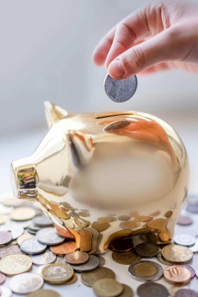 pexels-photo-12969339-12969339 Hand placing coin into a golden piggy bank surrounded by coins, symbolizing savings.